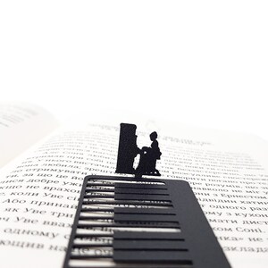 May include: A black metal bookmark featuring a piano keyboard design with a silhouette of a person playing the piano. The bookmark is placed on the pages of an open book, creating a musical theme.