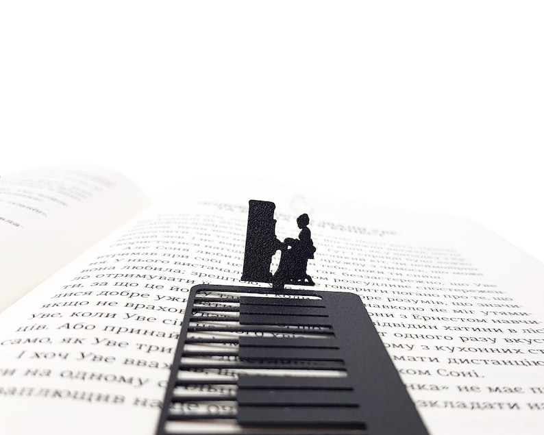 May include: A black metal bookmark featuring a piano keyboard design with a silhouette of a person playing the piano. The bookmark is placed on the pages of an open book, creating a musical theme.