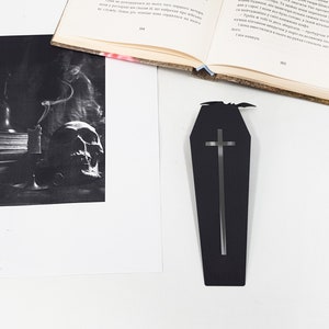 Black metal coffin-shaped bookmark with a cutout cross and small bat detail placed on a white surface beside an open book and a printed black-and-white photograph of a smoking candle and a skull.