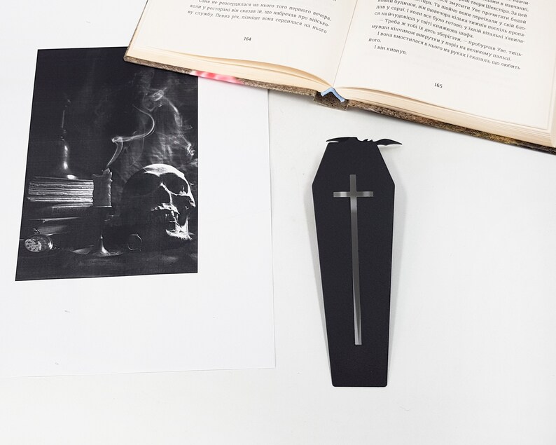 Black metal coffin-shaped bookmark with a cutout cross and small bat detail placed on a white surface beside an open book and a printed black-and-white photograph of a smoking candle and a skull.