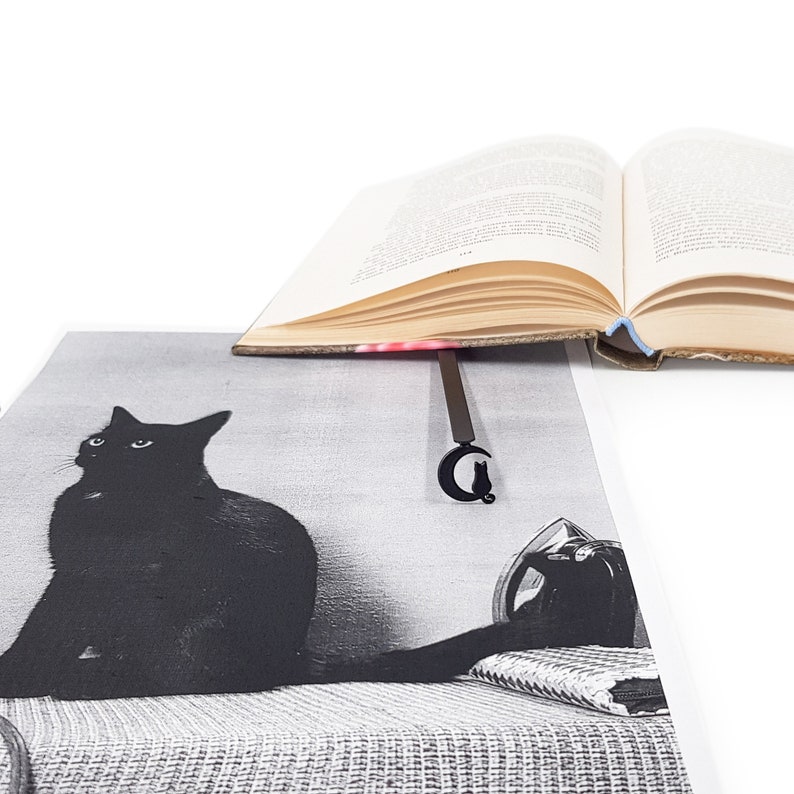 May include: A black cat sitting on a couch, looking to the left. A book with a black cat bookmark is on top of the couch.