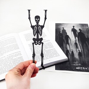 A hand holding a black metal skeleton-shaped bookmark in front of an open book, with a spooky black-and-white illustration of tall shadowy figures placed beside it.