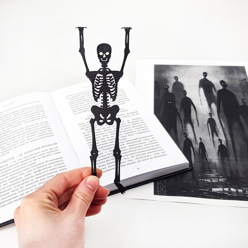 A hand holding a black metal skeleton-shaped bookmark in front of an open book, with a spooky black-and-white illustration of tall shadowy figures placed beside it.