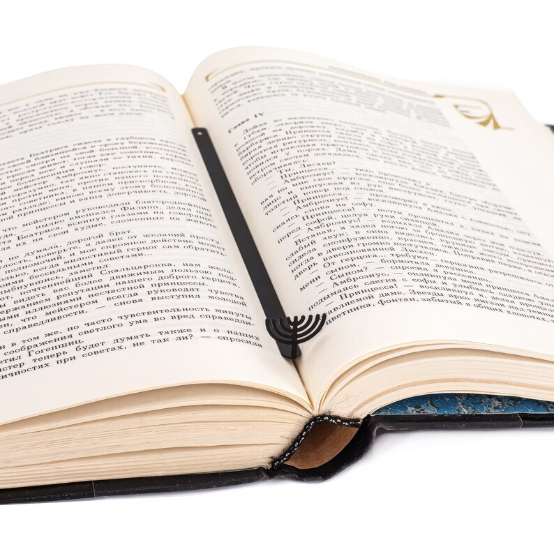 Black metal bookmark shaped like a menorah, resting neatly between the pages of an open book. A meaningful gift for Jewish readers and Hanukkah celebrations.