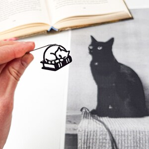 Op de afbeelding: Zwart metalen boekenlegger met een slapende kat op boeken. De boekenlegger wordt door een hand vastgehouden, met een zwart-wit foto van een zwarte kat op de achtergrond. Een open boek is zichtbaar.