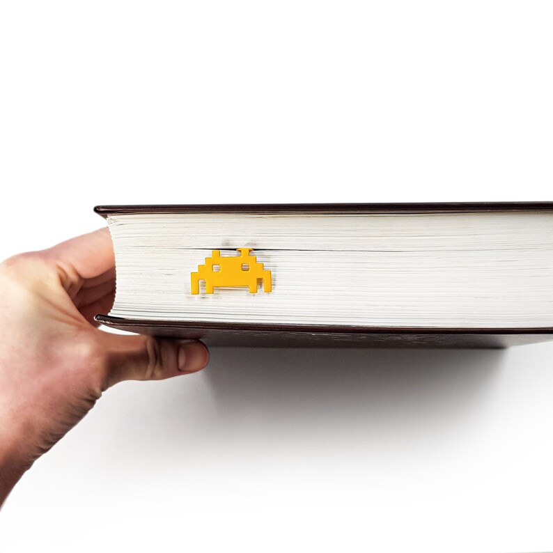 May include: A yellow plastic bookmark shaped like a classic video game alien. The bookmark is placed on a closed hardcover book with a brown cover.