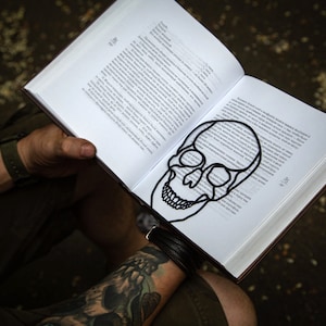 Minimalist style black metal bookmark. The face of a Skul is placed between the pages of the book. Cross bones are seen on the side of the book even when the book is closed.