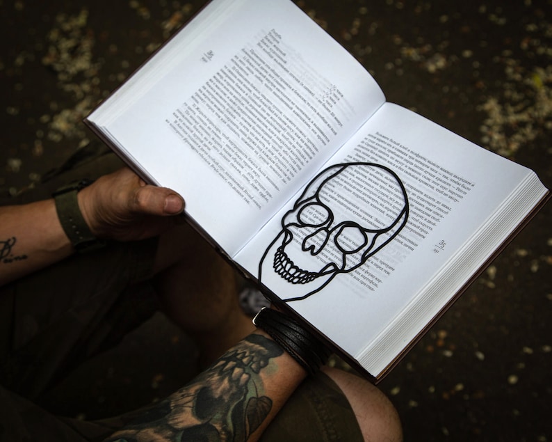 Minimalist style black metal bookmark. The face of a Skul is placed between the pages of the book. Cross bones are seen on the side of the book even when the book is closed.