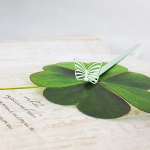 May include: A light green butterfly bookmark with a four-leaf clover design. The bookmark is made of paper and has a delicate, intricate design.
