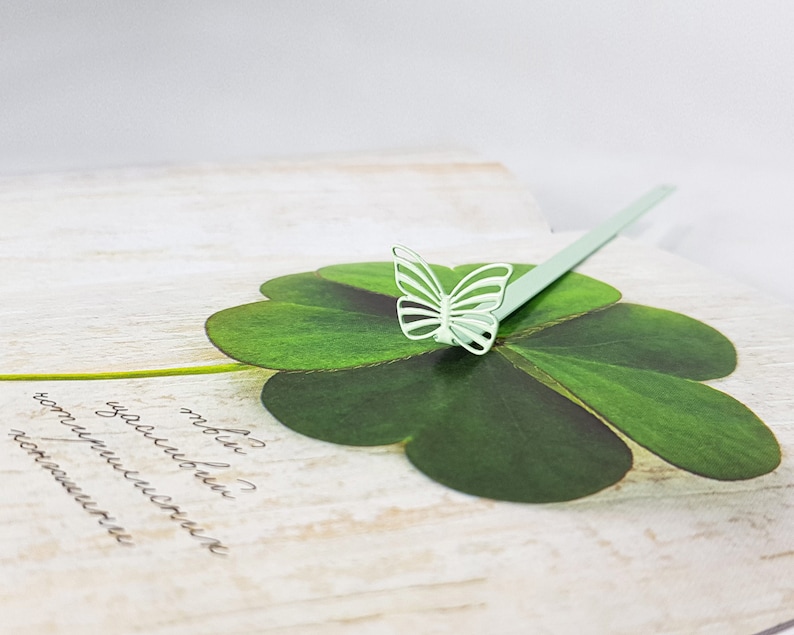 May include: A light green butterfly bookmark with a four-leaf clover design. The bookmark is made of paper and has a delicate, intricate design.
