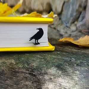 Black metal raven-shaped bookmark peeking from a yellow-edged book, resting on a mossy surface with autumn leaves. A moody fall reading gift with gothic charm.