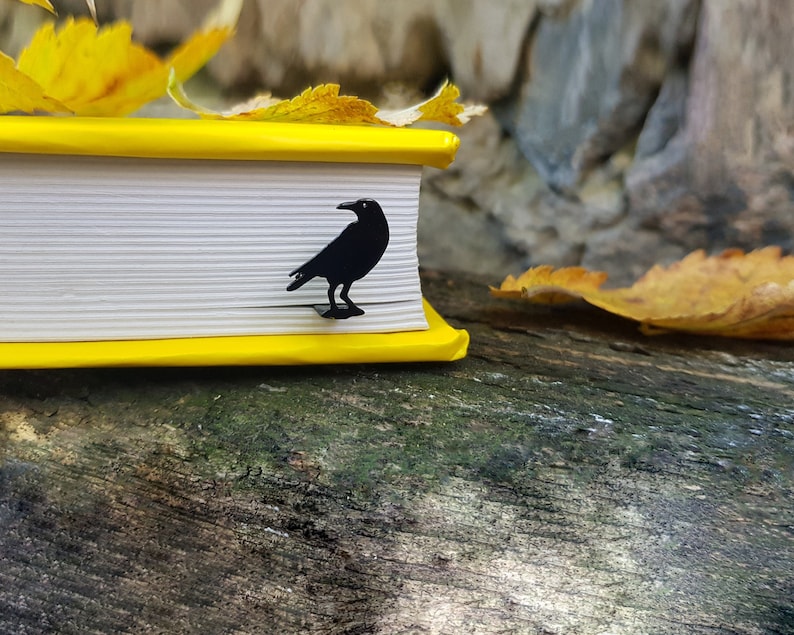 Black metal raven-shaped bookmark peeking from a yellow-edged book, resting on a mossy surface with autumn leaves. A moody fall reading gift with gothic charm.