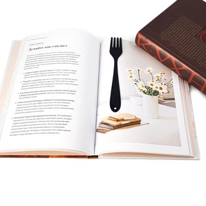 May include: An open book with a black bookmark shaped like a fork. The book is open to a page with text in Russian. The book is lying on a wooden surface.