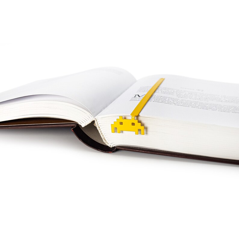 May include: A yellow metal bookmark shaped like a retro video game alien. The bookmark is placed in a book with the pages open.