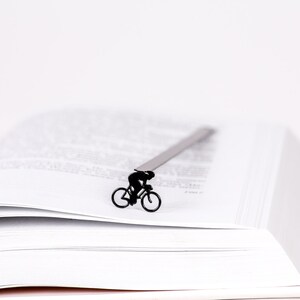 A black metal bookmark shaped like a cyclist riding a bicycle. The bookmark is placed on a white book with pages turned over.