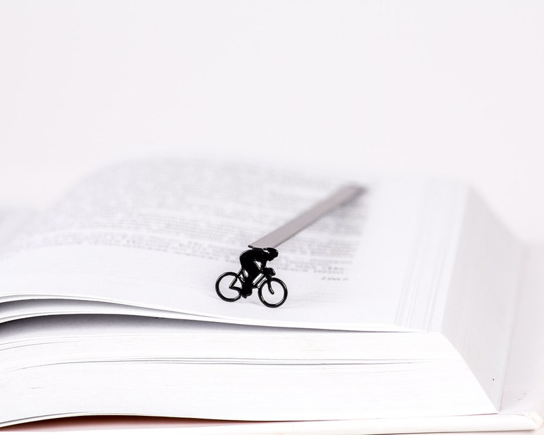 A black metal bookmark shaped like a cyclist riding a bicycle. The bookmark is placed on a white book with pages turned over.