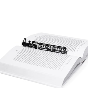 Może przedstawiać: Czarny metalowy zakładka w kształcie pociągu z napisem "READ" z boku. Zakładka znajduje się w otwartej książce.
