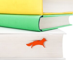 May include: A stack of three books with a white cover, a green cover, and a yellow cover. A red fox bookmark is sticking out of the top book.