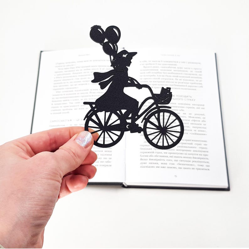 Hand holding a black metal bike bookmark shaped as a girl riding a bicycle with balloons, positioned in front of an open book on a white background.
