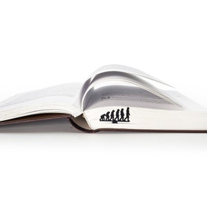May include: A black metal bookmark shaped like the evolution of humans, from ape to modern human, is placed in an open book.