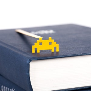 May include: A yellow space invader bookmark on a dark blue book. The bookmark is made of metal and has a gold finish.