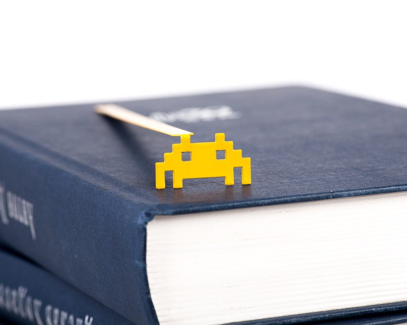 May include: A yellow space invader bookmark on a dark blue book. The bookmark is made of metal and has a gold finish.