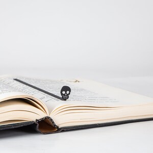 May include: A black skull bookmark is placed in a book with white pages. The bookmark is made of metal and has a simple design.