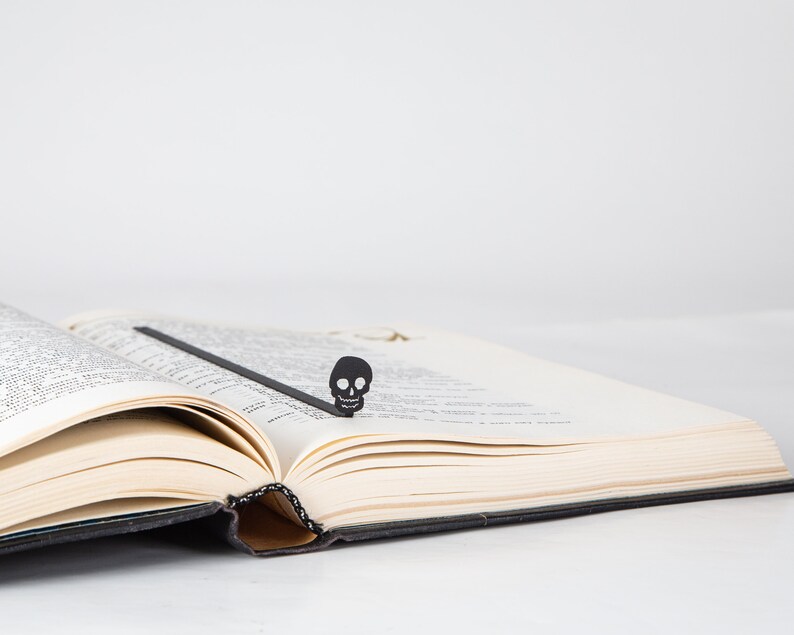 May include: A black skull bookmark is placed in a book with white pages. The bookmark is made of metal and has a simple design.