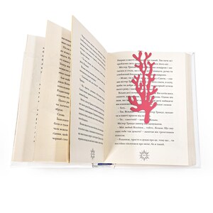 May include: A red metal bookmark shaped like a coral branch. The bookmark is placed in an open book with text in a foreign language.