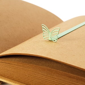 May include: A brown leather-bound journal with a green metal butterfly bookmark. The bookmark is attached to the journal with a thin metal ribbon.