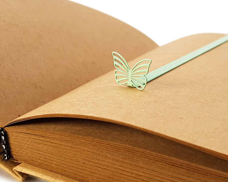 May include: A brown leather-bound journal with a green metal butterfly bookmark. The bookmark is attached to the journal with a thin metal ribbon.