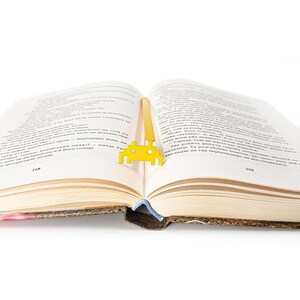 May include: A yellow metal bookmark shaped like a retro video game alien. The bookmark is placed between the pages of an open book.