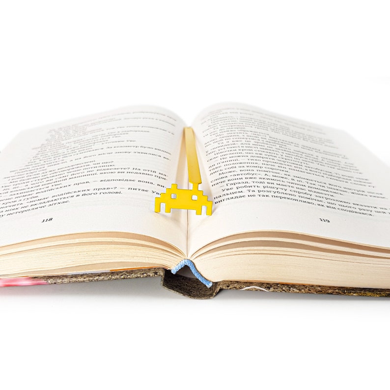 May include: A yellow metal bookmark shaped like a retro video game alien. The bookmark is placed between the pages of an open book.