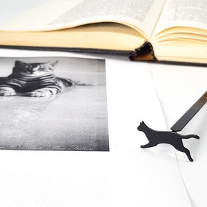 Black metal running cat bookmark lying beside an open book and a black-and-white photograph of a resting cat.