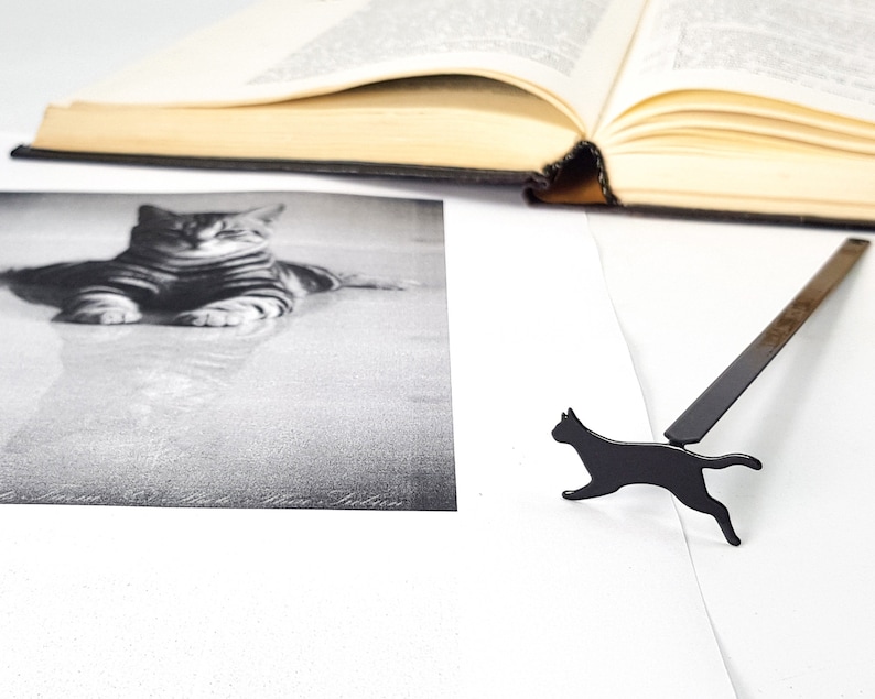 Black metal running cat bookmark lying beside an open book and a black-and-white photograph of a resting cat.