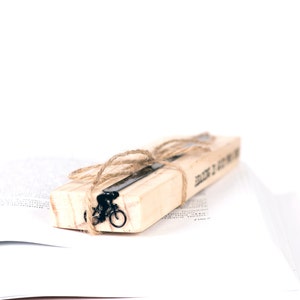 A wooden packaging block with a black metal bookmark with bicycle design and the words 'Read It, Got It, Done' printed on it. The bookmark is tied with twine and is resting on a white book.