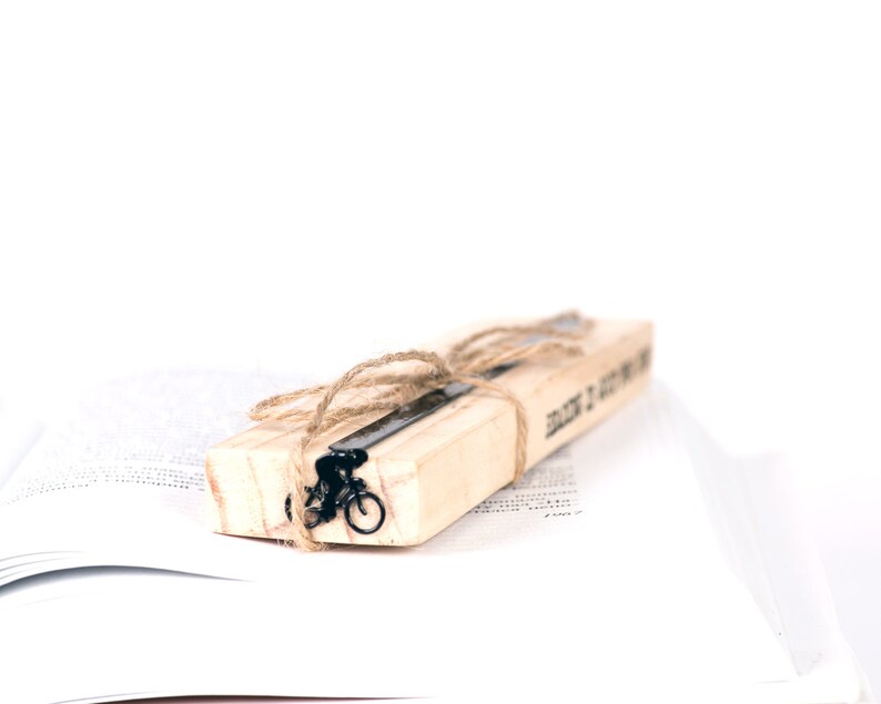 A wooden packaging block with a black metal bookmark with bicycle design and the words 'Read It, Got It, Done' printed on it. The bookmark is tied with twine and is resting on a white book.