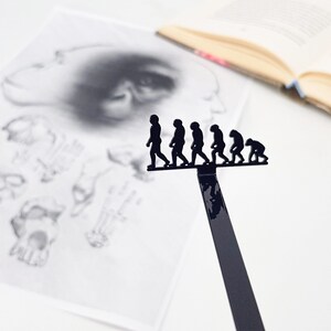 May include: A black metal bookmark shaped like a line of figures representing the evolution of humans. The figures are in silhouette and are walking in a line from left to right.