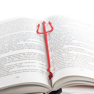 Puede incluir: Un marcapáginas de metal rojo con forma de tridente, una lanza de tres puntas, está colocado en un libro abierto. El marcapáginas es una forma divertida y única de marcar tu lugar en un libro.