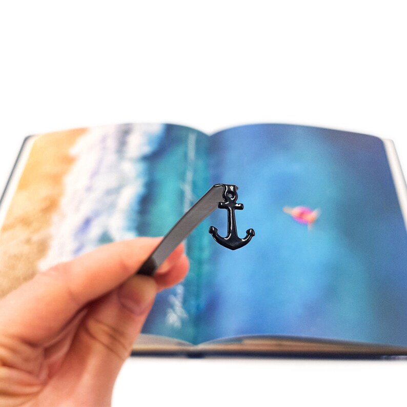 May include: A black metal bookmark shaped like an anchor, with a black ribbon attached. The bookmark is being held over an open book with a watercolor painting of a beach scene.
