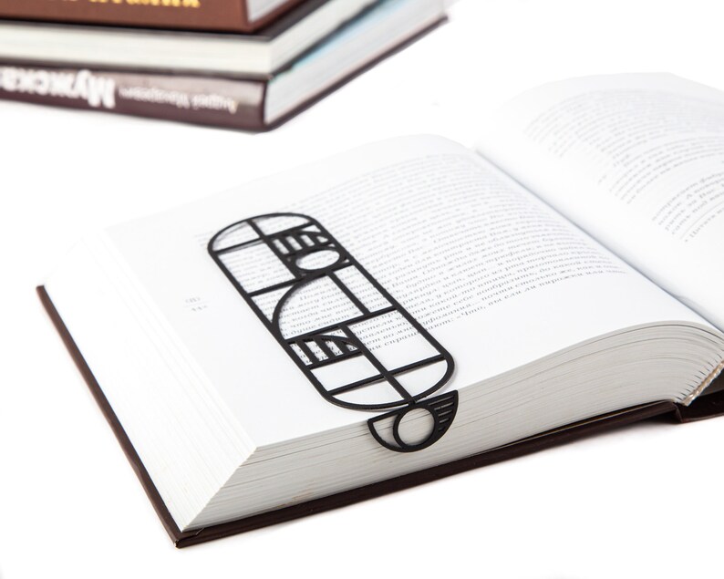 A black metal bookmark Bauhaus in the shape of a geometric design. The bookmark is placed in a book with white pages. Not mass-produced.