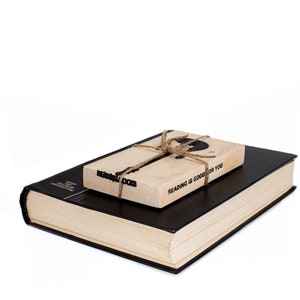 Black metal guitar-shaped bookmark with the phrase READING ROCKS, tied to a wooden block that says Reading is good for you, resting on a closed book. A fun gift for readers who love rock music.