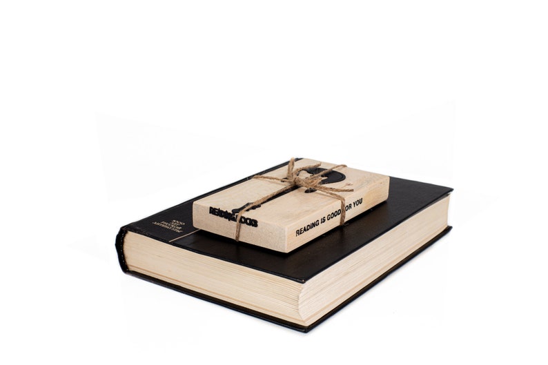 Black metal guitar-shaped bookmark with the phrase READING ROCKS, tied to a wooden block that says Reading is good for you, resting on a closed book. A fun gift for readers who love rock music.