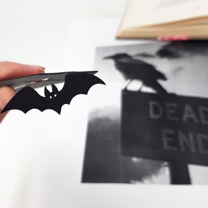 Black metal bat detail from a coffin-shaped bookmark held in hand, shown in front of a black-and-white image of a crow perched on a “DEAD END” sign, with an open book blurred in the background.