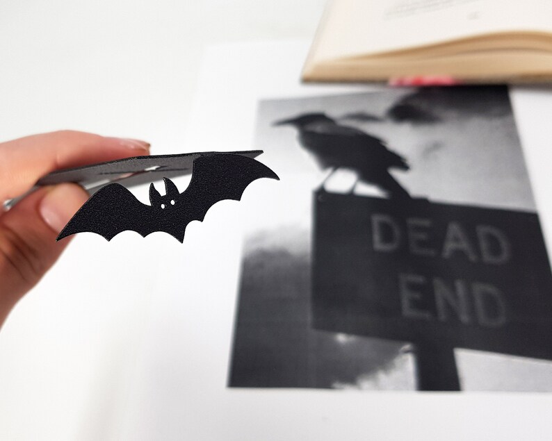 Black metal bat detail from a coffin-shaped bookmark held in hand, shown in front of a black-and-white image of a crow perched on a “DEAD END” sign, with an open book blurred in the background.