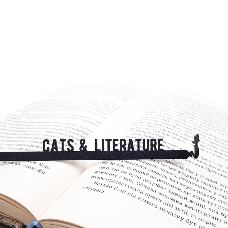 Black metal bookmark Cats and Literature. A flat metal stick stays between the pages of the book, the writing Cats and Literature is on the stick. A silhouette of the Laying Cat is on the side of the book. It is visible even when the book is closed.