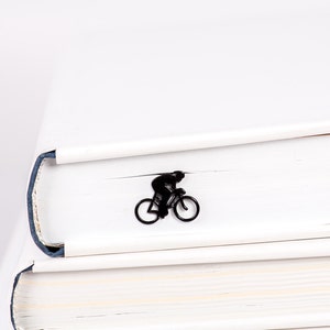 A black minimalis style metal bookmark shaped like a person riding a bicycle. The bookmark is inside a book.