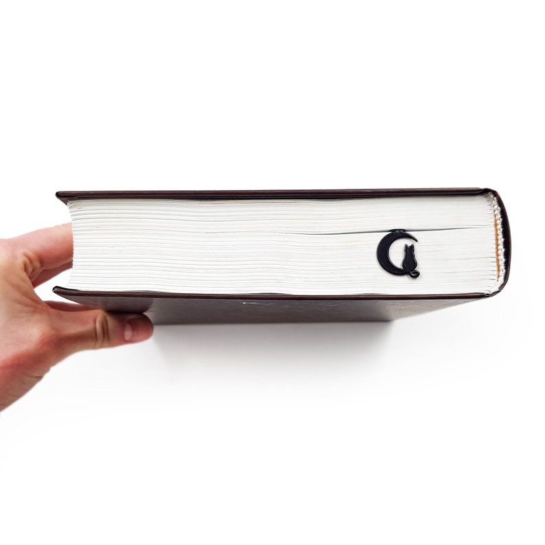 May include: A close-up of a thick, brown book with a black cat and crescent moon bookmark. The book is held in a hand.