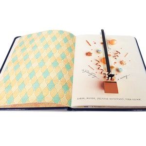 May include: Open notebook with a blue cover and patterned pages. The left page has a yellow and teal diamond pattern. The right page has a black cat bookmark with a ribbon and decorative elements. Text in the image reads: "Wish, love, save, photograph, make collages!"