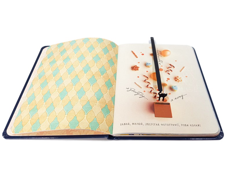 May include: Open notebook with a blue cover and patterned pages. The left page has a yellow and teal diamond pattern. The right page has a black cat bookmark with a ribbon and decorative elements. Text in the image reads: "Wish, love, save, photograph, make collages!"